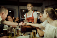 Servicekraft in Tracht serviert am Tisch mehrere Schnapsgläser an eine Gästegruppe in der Hoftaverne